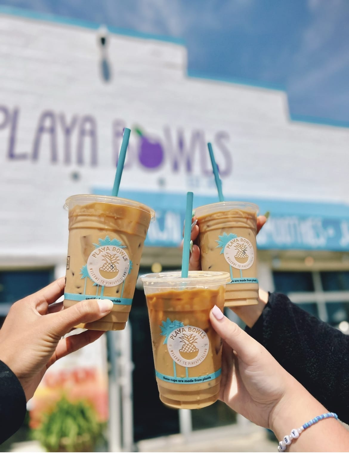 Three Playa Bowls plastic cups with blue straws containing cold brew coffee with a Playa Bowls store in the background